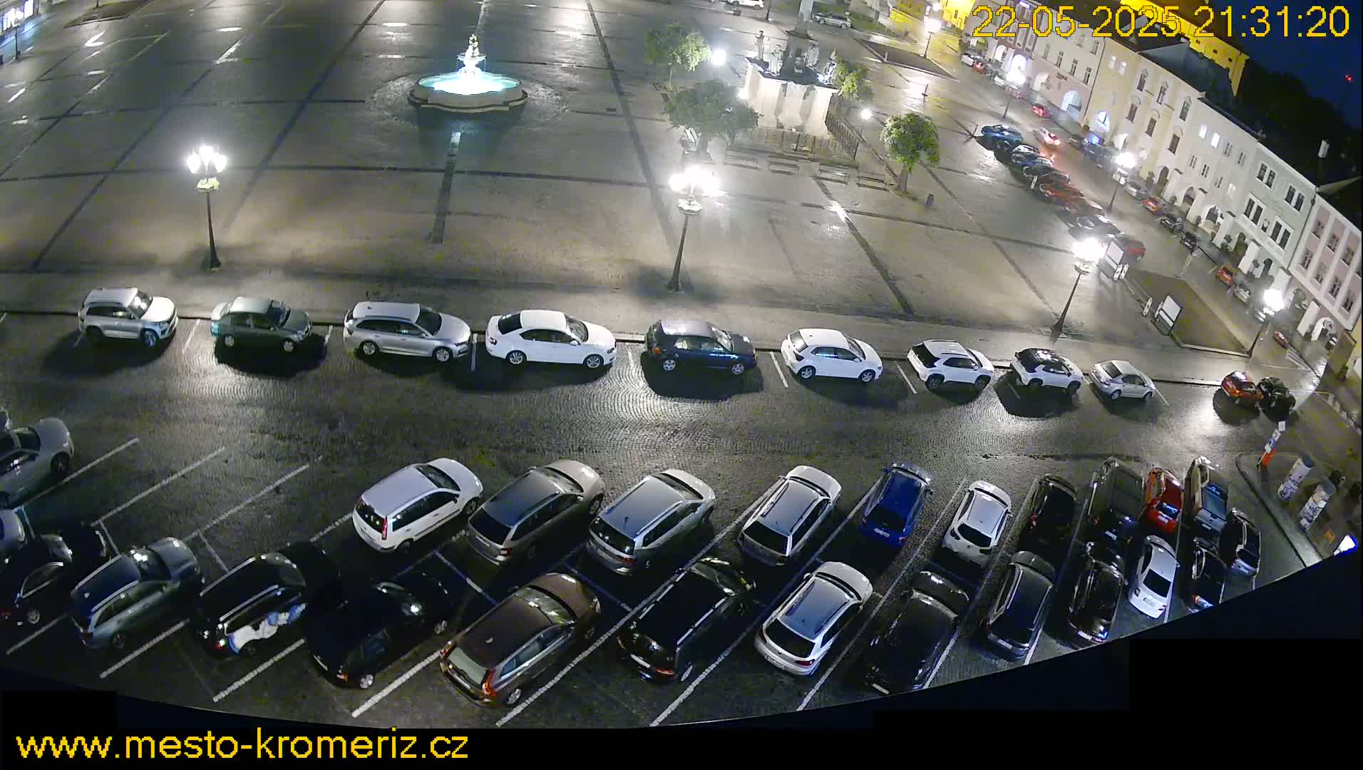 A nighttime, high-angle view of a town square showing a fountain, several parked cars, and a few cars driving on the street.
