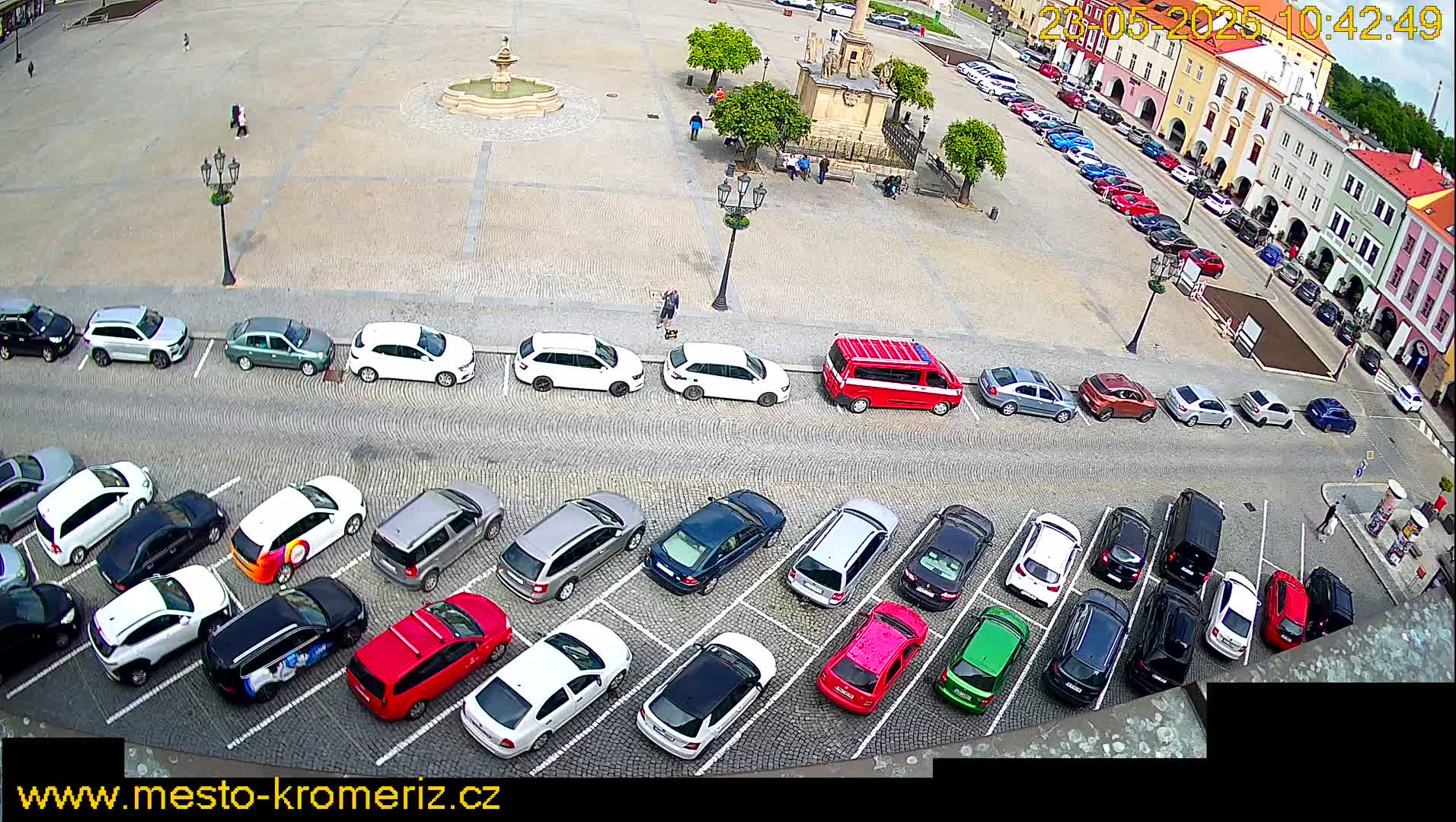 A town square with a fountain and numerous cars parked in rows, under a sunny sky.