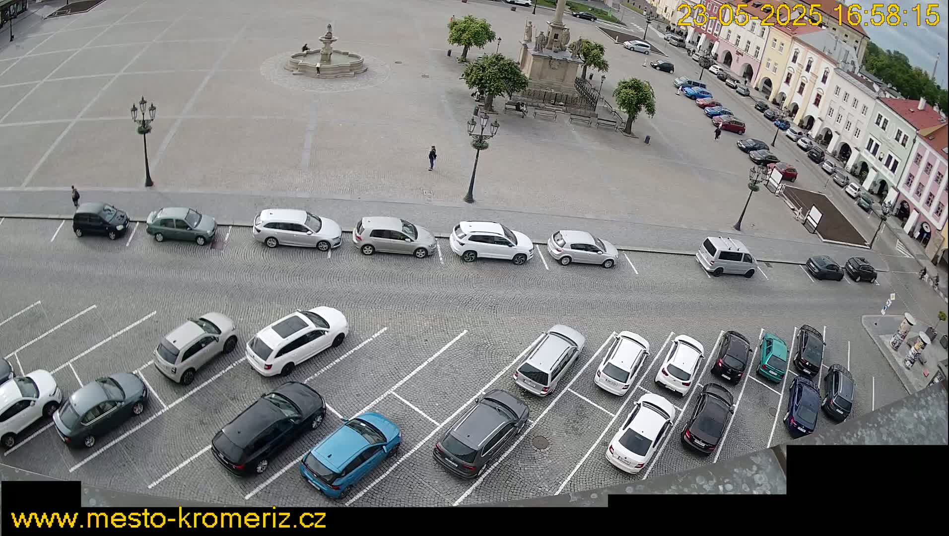 A paved square with a fountain and several parked cars is viewed from above on an overcast day.