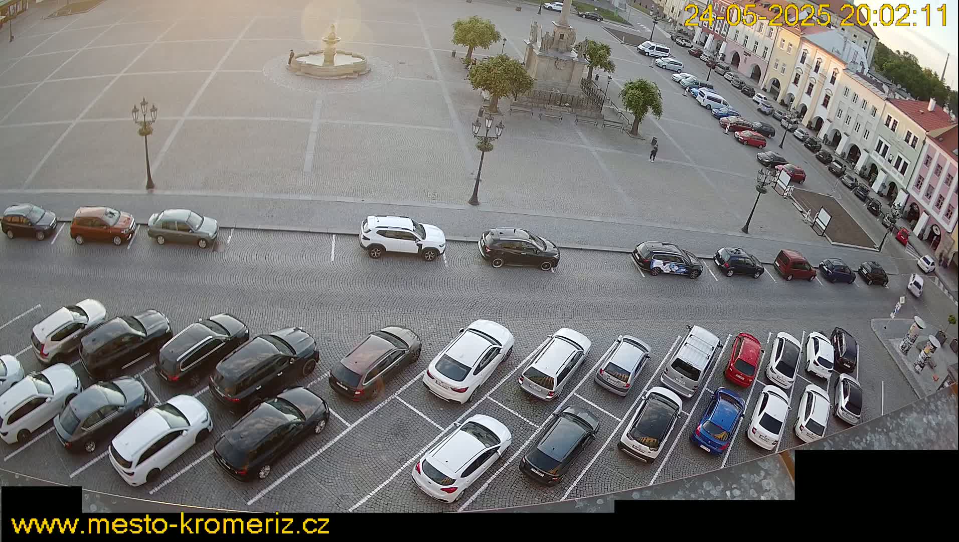 A cobblestone town square, under a partly sunny sky, contains a fountain and numerous cars parked in rows.