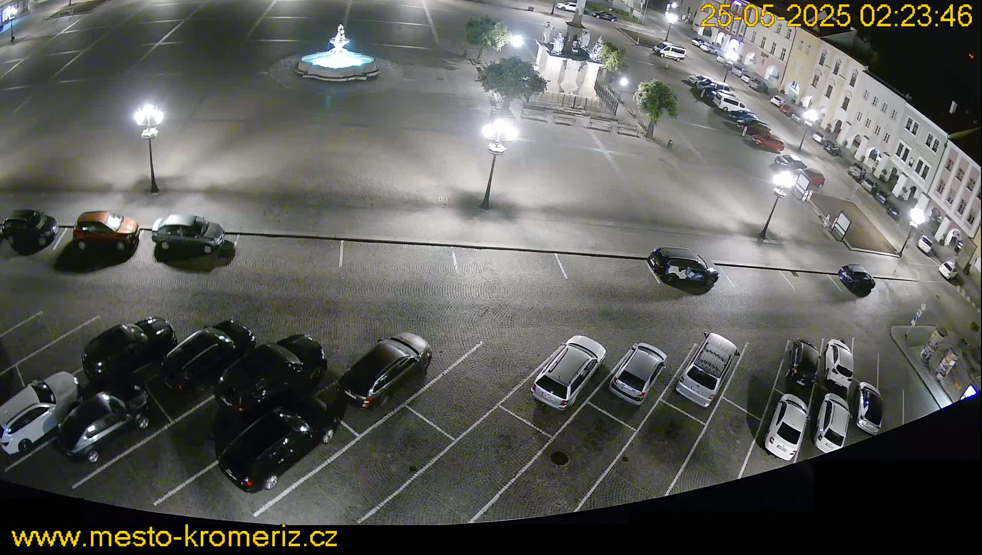 A nighttime view of a town square with a fountain, several cars parked in a lot, and streetlights illuminating the scene.