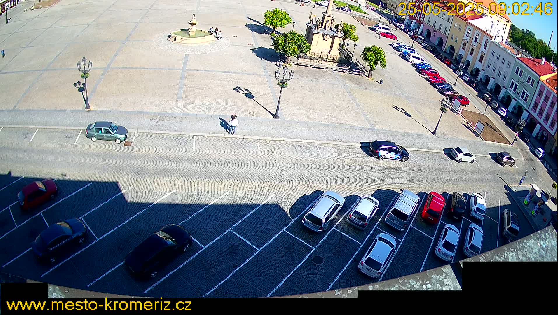 A sunny day reveals a town square with a fountain, several parked cars, and a few pedestrians; nearby, a parking lot is also filled with vehicles.