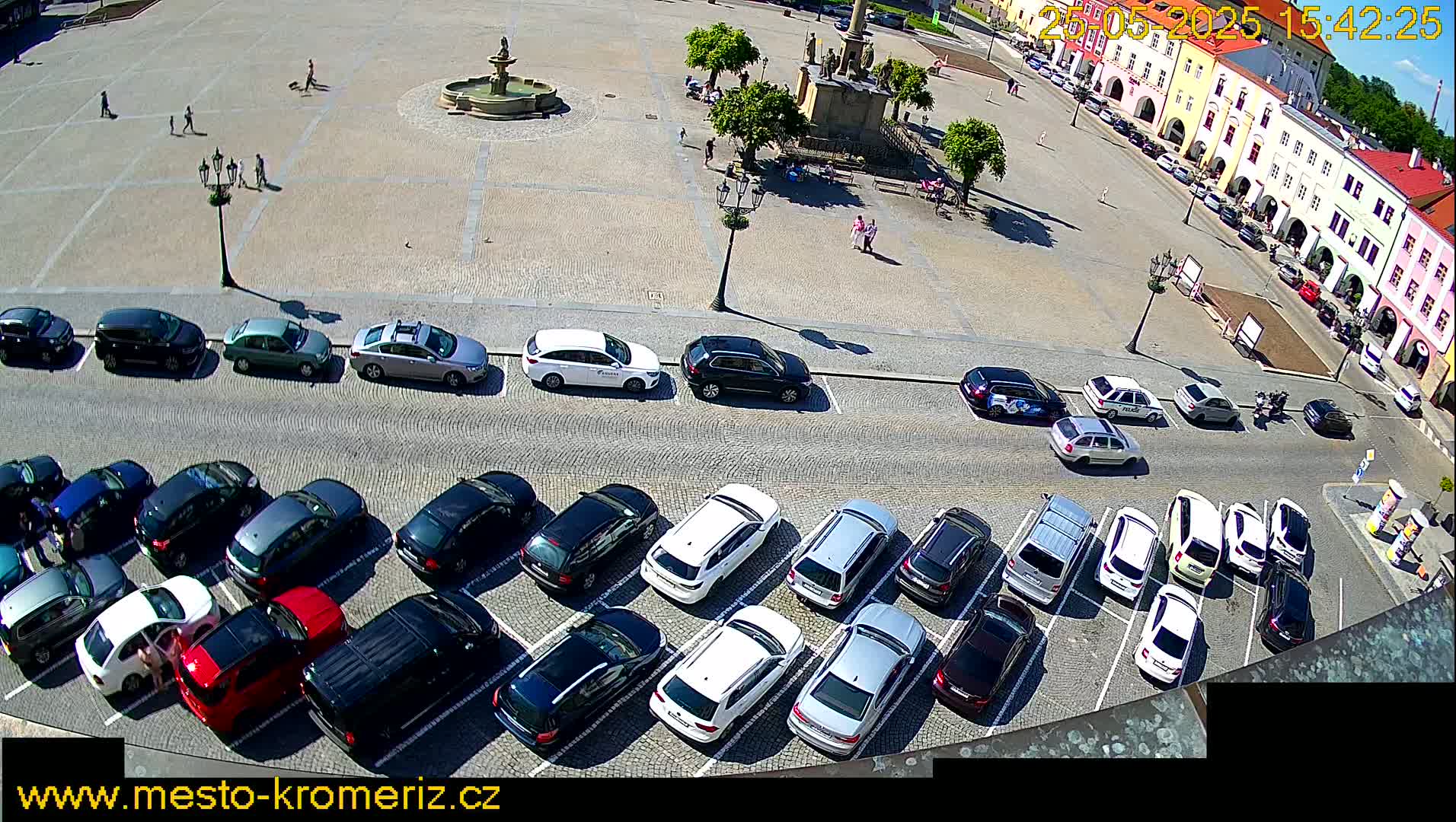 A sunny day shows a town square with a fountain and several parked cars lining the perimeter, while a few people stroll across the plaza.