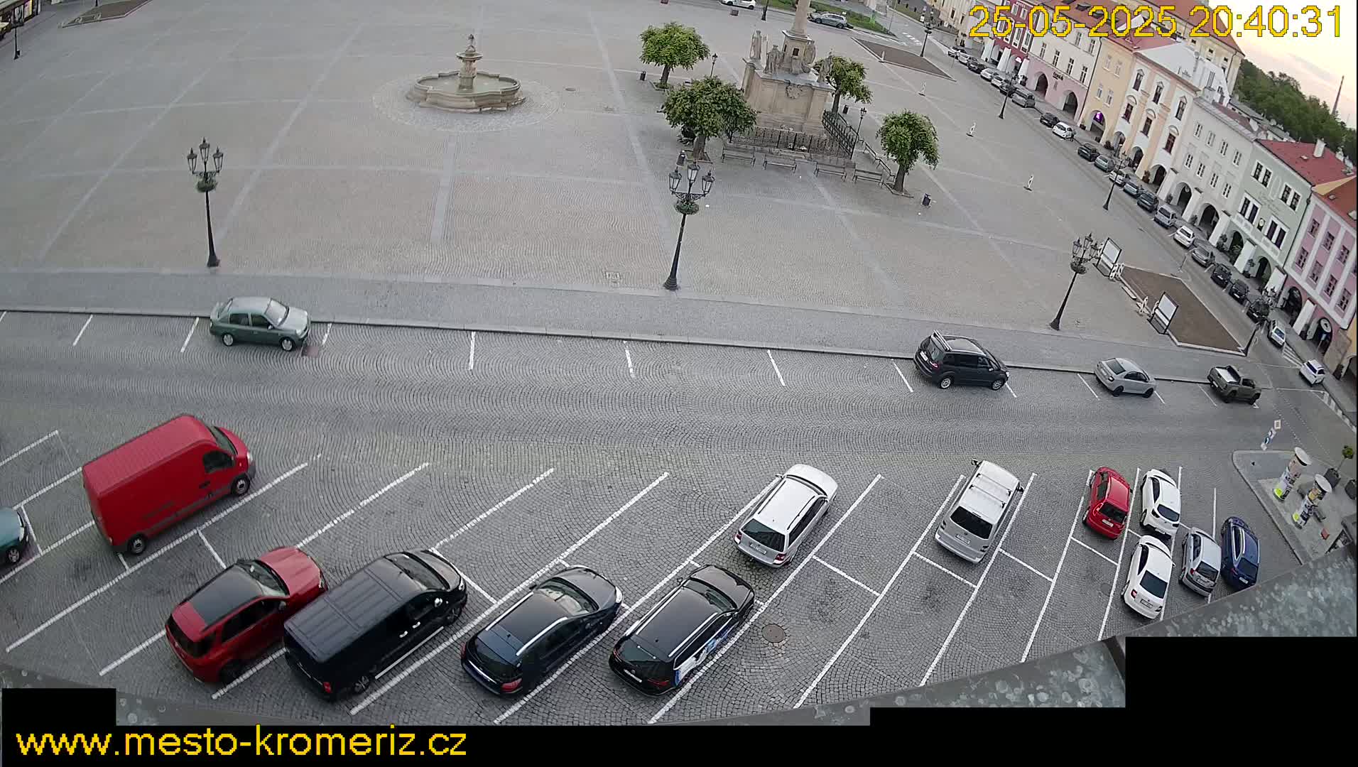 An aerial view of a town square with a fountain and several parked cars in a cobblestone parking lot under a partly cloudy sky.