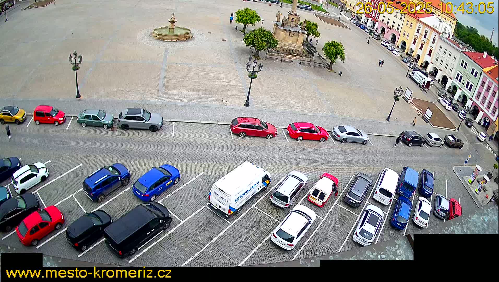 An aerial view of a town square with a fountain and many cars parked in a lot next to a row of buildings under partly cloudy skies.