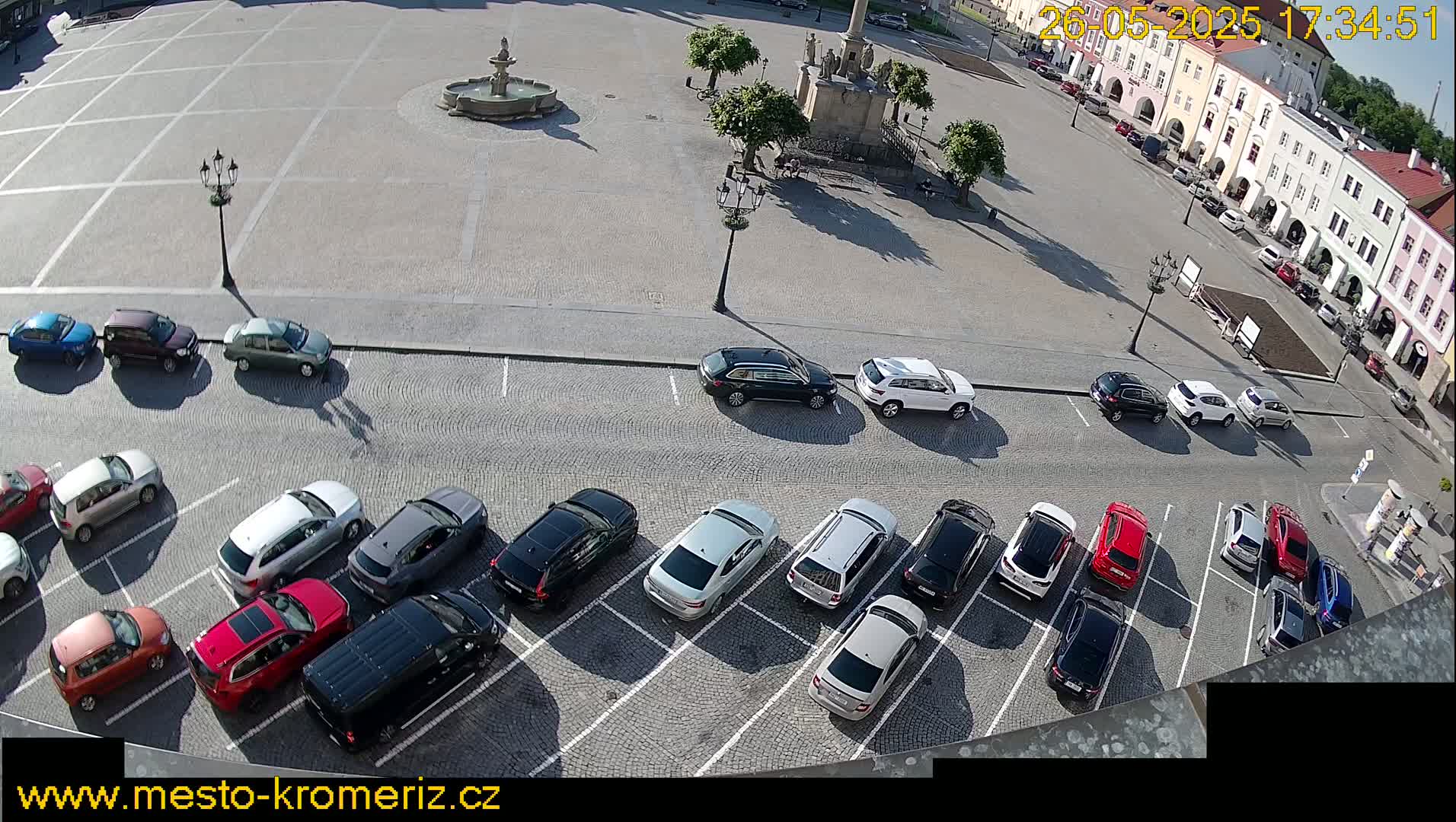 A sunny day reveals a town square with a fountain, surrounded by parked cars and buildings.