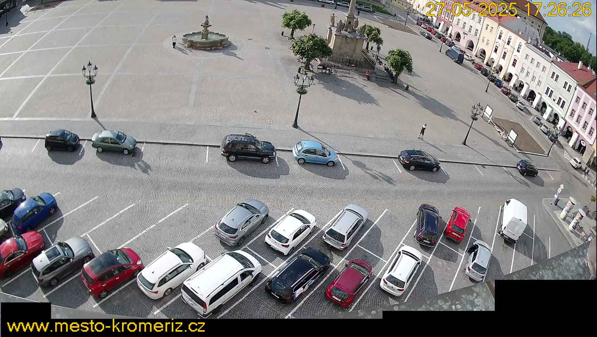 A town square with a fountain, surrounded by buildings and numerous cars parked in a lot adjacent to the square on a sunny day.