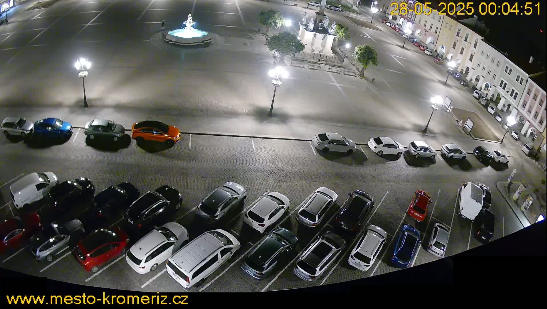A nighttime aerial view of a town square with a fountain, several streetlights, and a parking lot full of cars.