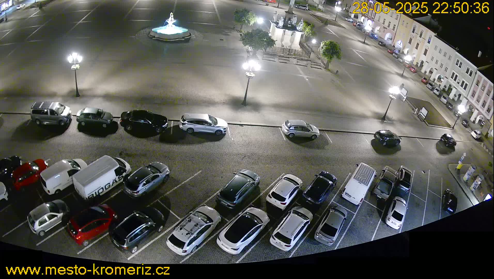 A nighttime aerial view of a town square with a fountain at its center, surrounded by parked cars under artificial lighting.