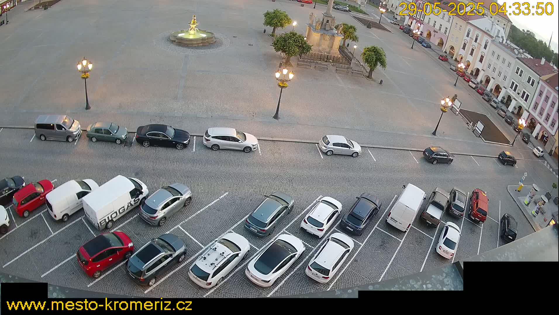 A cobblestone town square with a fountain and numerous cars parked in a lot, under partly cloudy evening conditions.