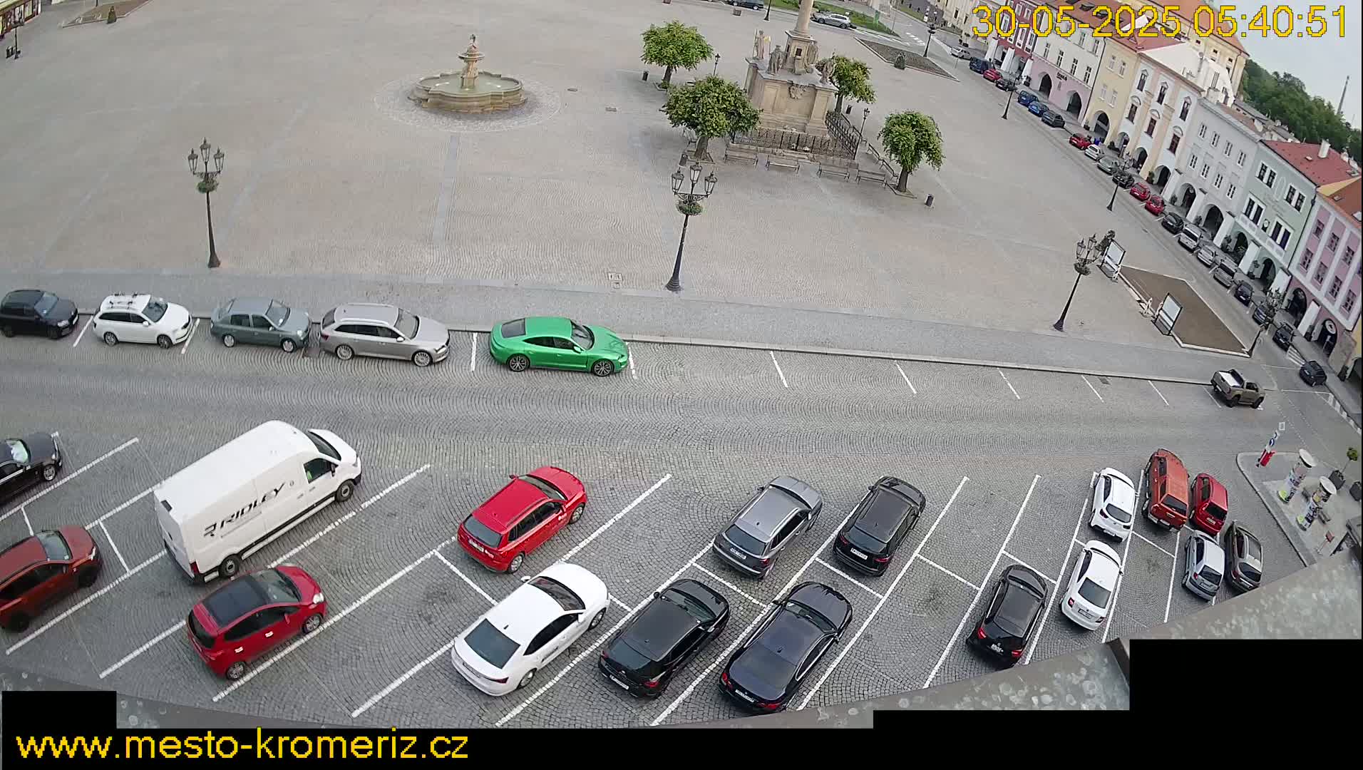 A high-angle view of a town square with parked cars, a fountain, and several buildings surrounding the square on a partly sunny day.