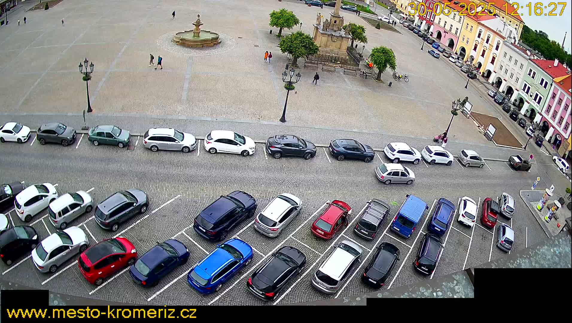 A town square with a fountain and several parked cars is viewed from above on an overcast day.