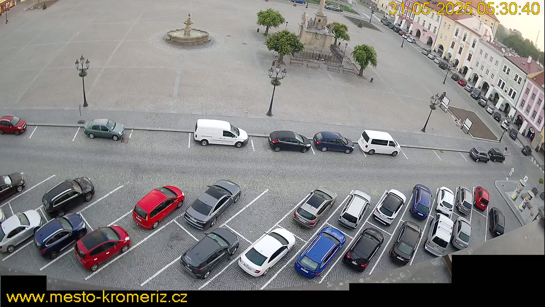 A town square with a fountain and several parked cars is viewed from above on a partly cloudy day.