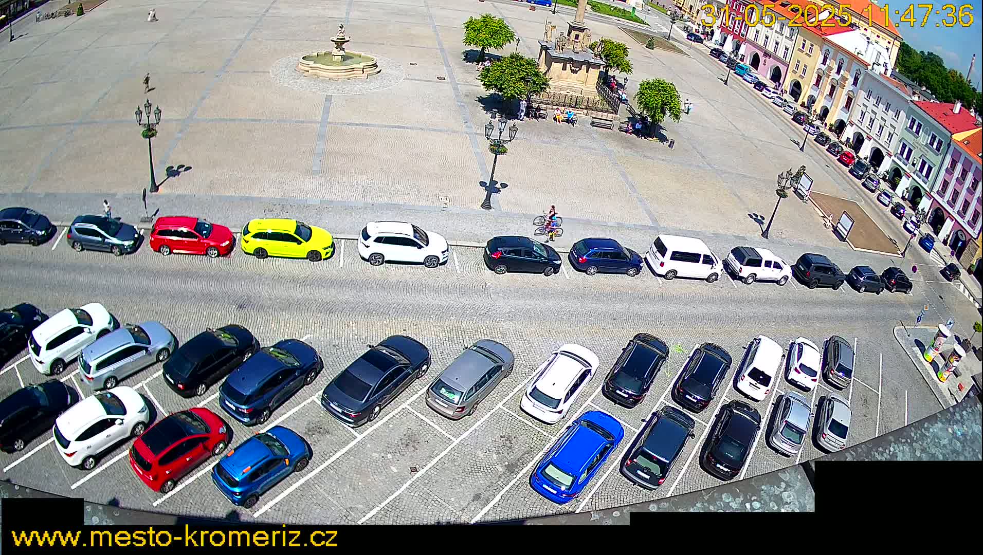 A sunny day reveals a town square with a fountain and many cars parked in rows on a cobblestone surface.