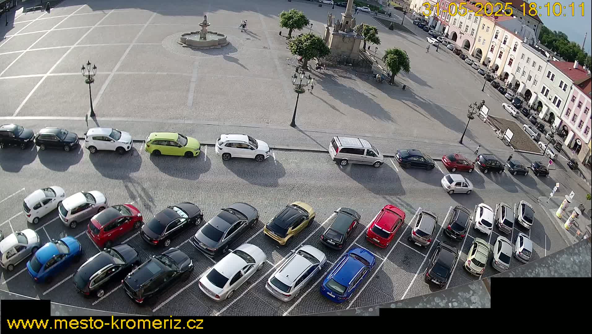 A paved town square with a fountain in the center, surrounded by buildings and numerous cars parked in rows.
