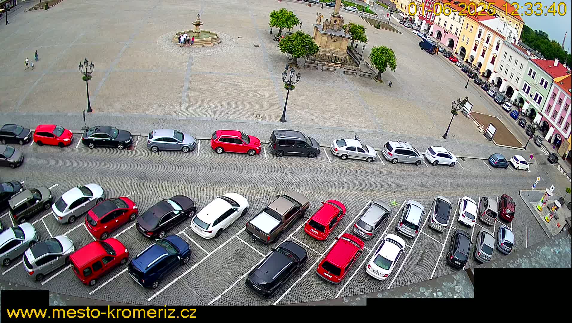 A cobblestone town square with a fountain and a monument, surrounded by parked cars, under a partly cloudy sky.