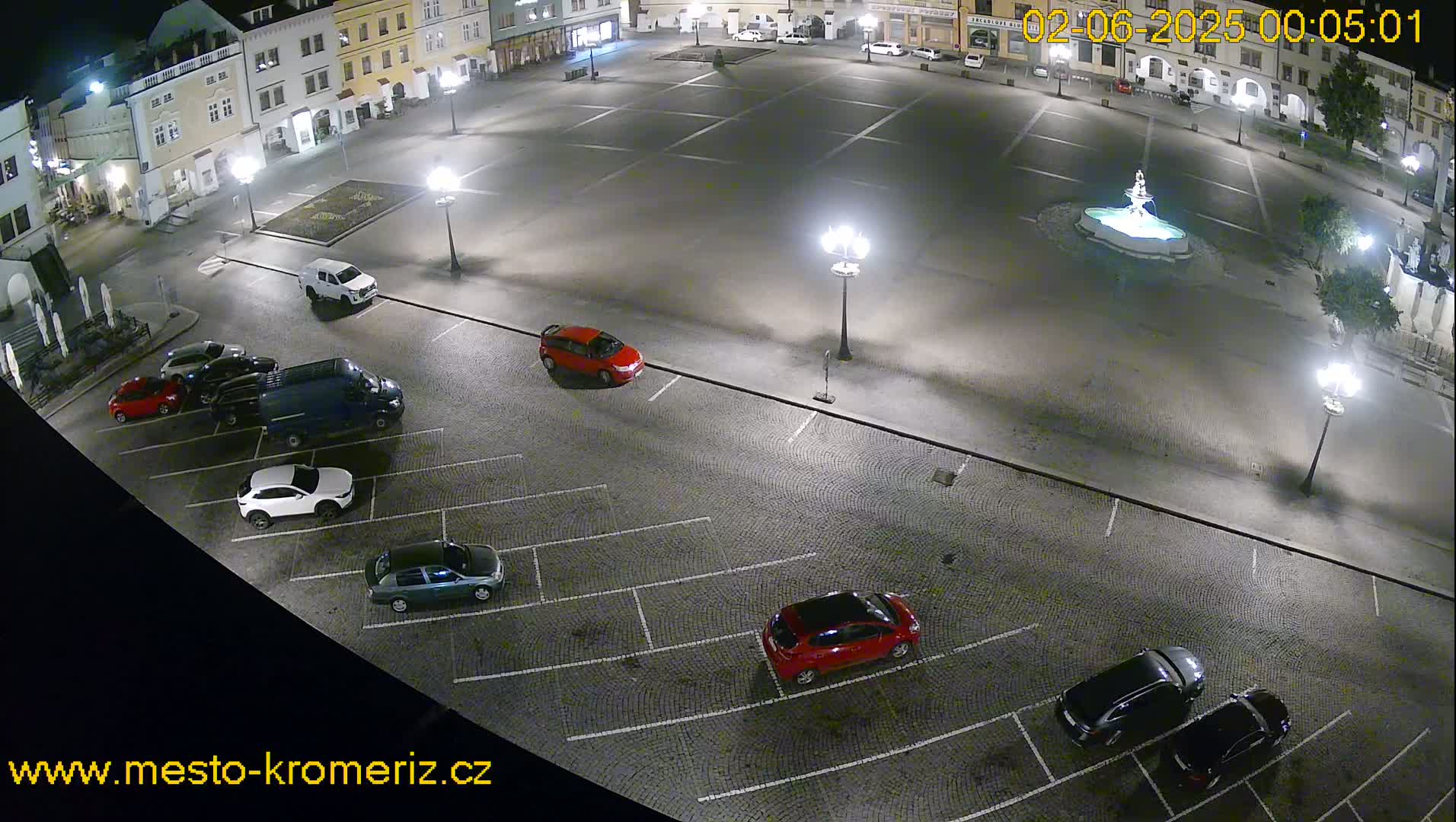 A nighttime aerial view of a town square with a fountain at its center and several cars parked in a surrounding lot.