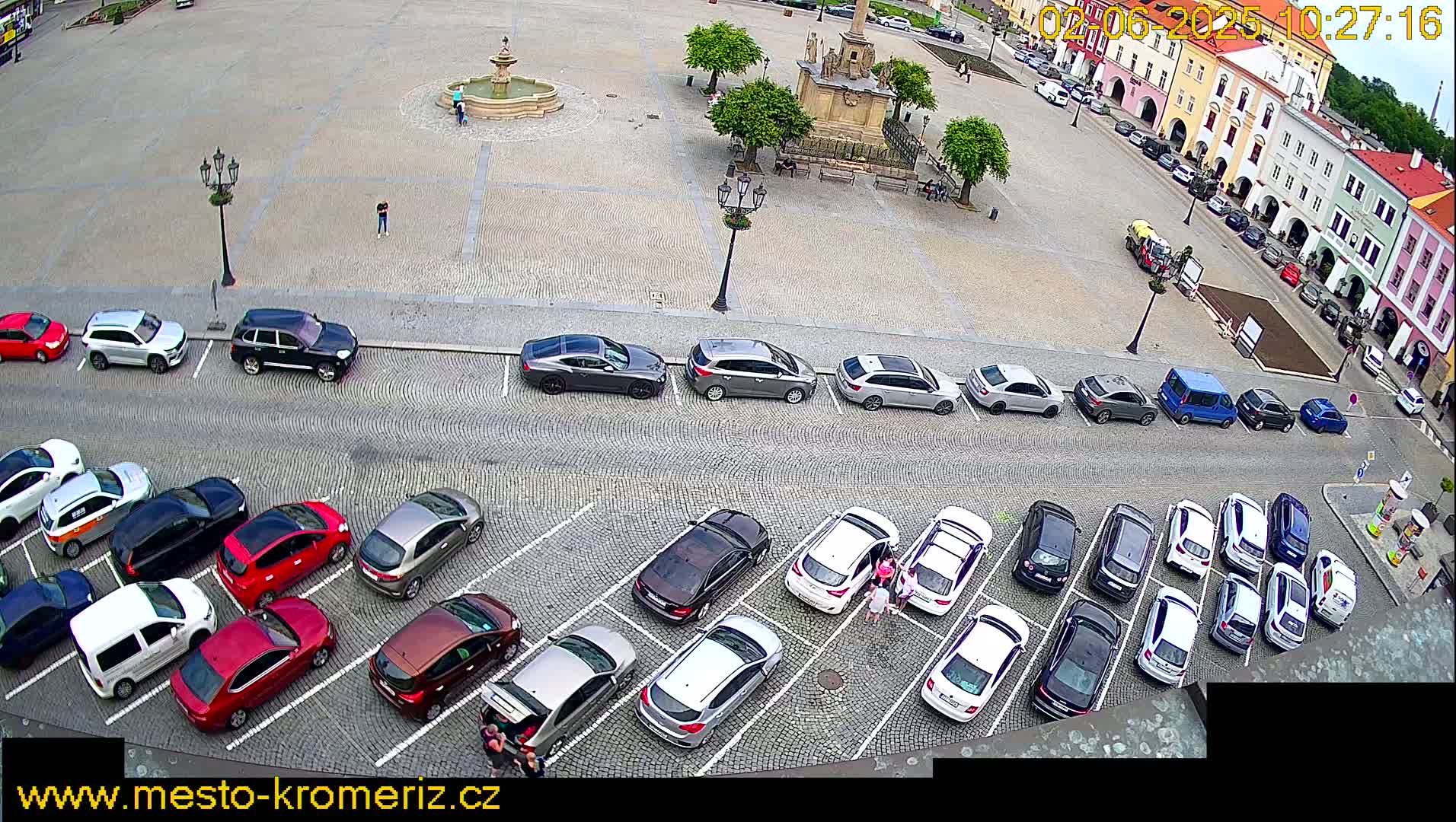 A town square with a fountain and several cars parked in the surrounding lots on a partly cloudy day.
