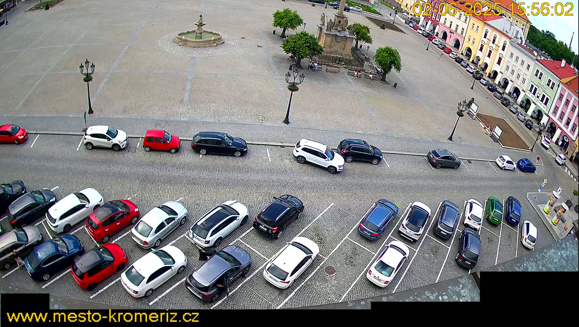 A high-angle view shows a town square with a fountain and several cars parked in the surrounding area on a mostly sunny day.