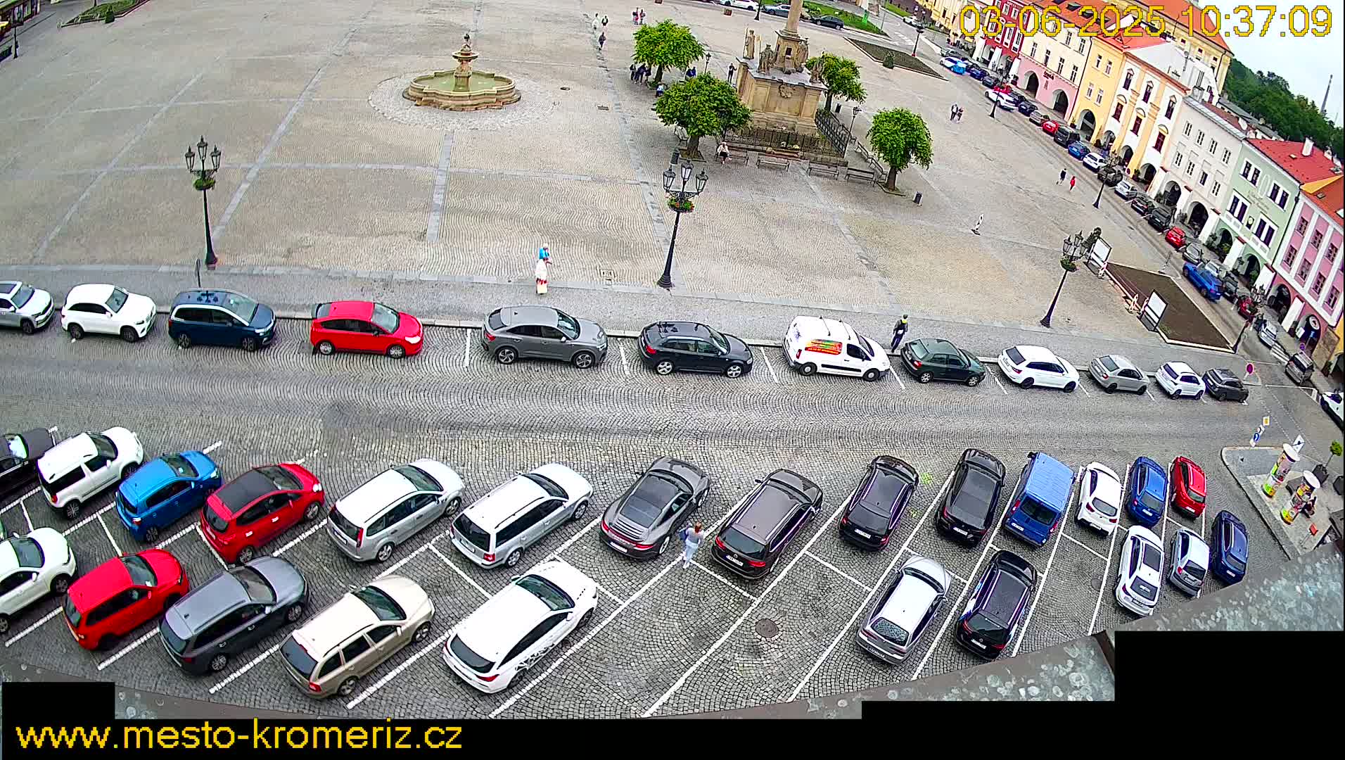 A high-angle view shows a town square with a fountain and several cars parked in rows, under an overcast sky.