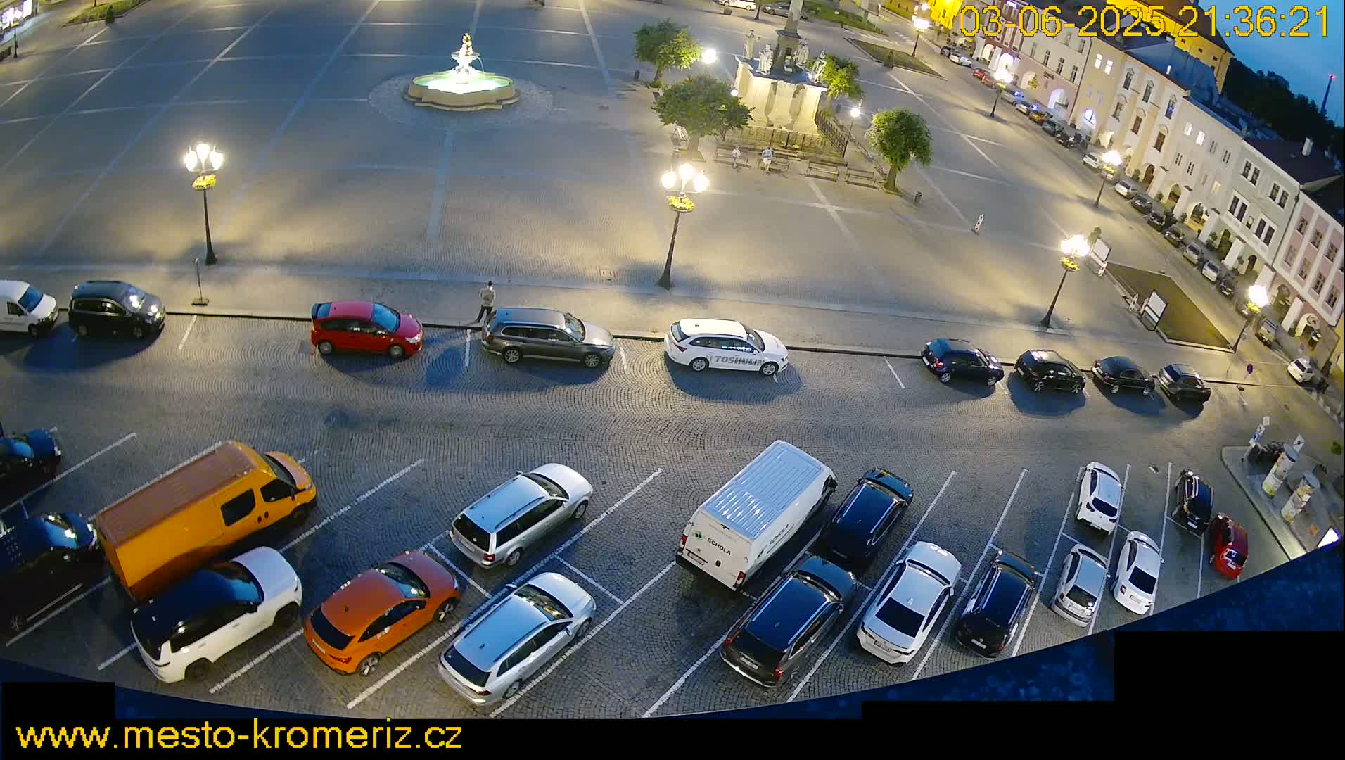 A nighttime aerial view of a town square parking lot with numerous cars parked and a fountain in the center, under partly cloudy skies.