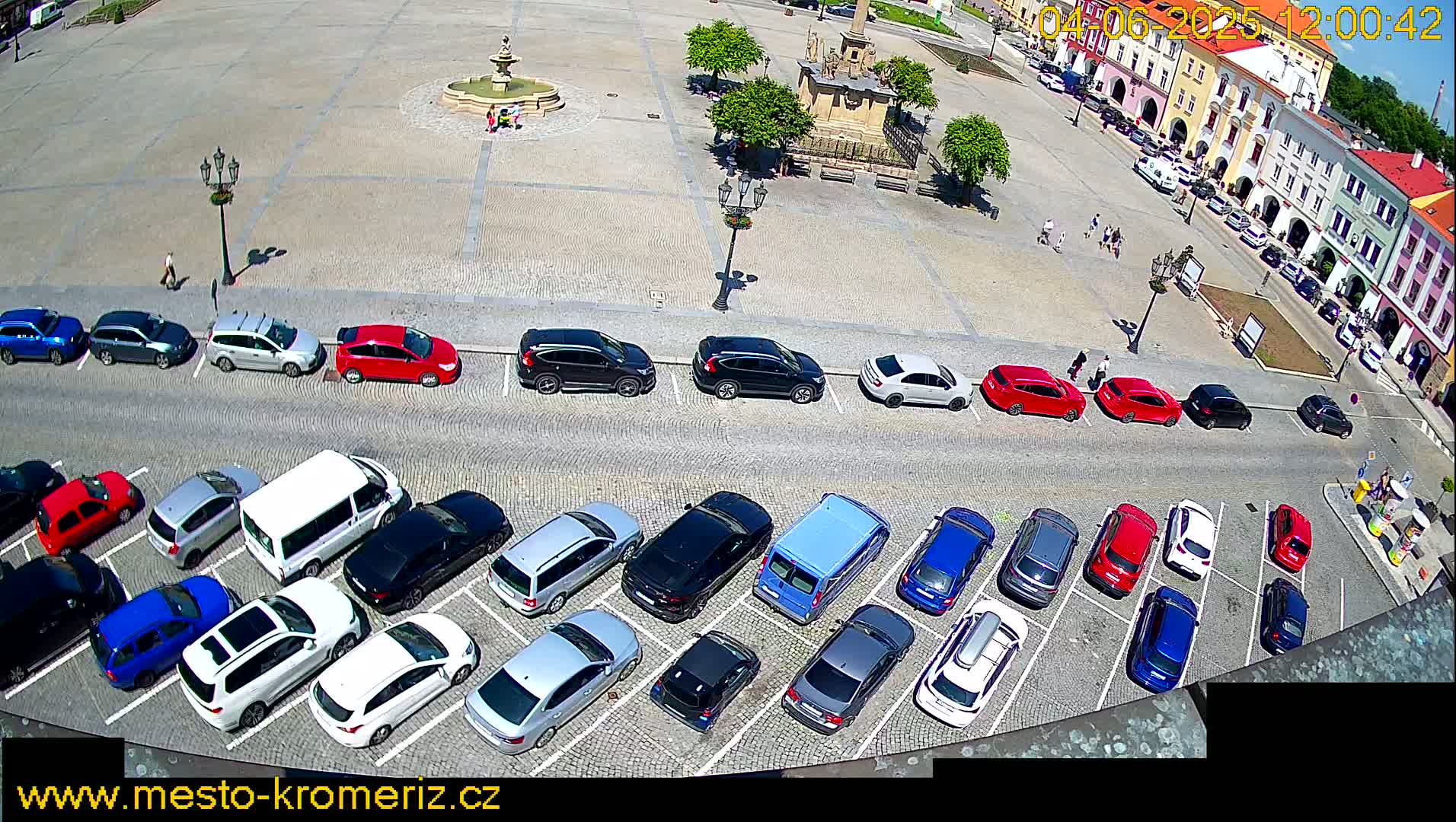 A parking lot filled with various cars is situated next to a town square on a sunny day.