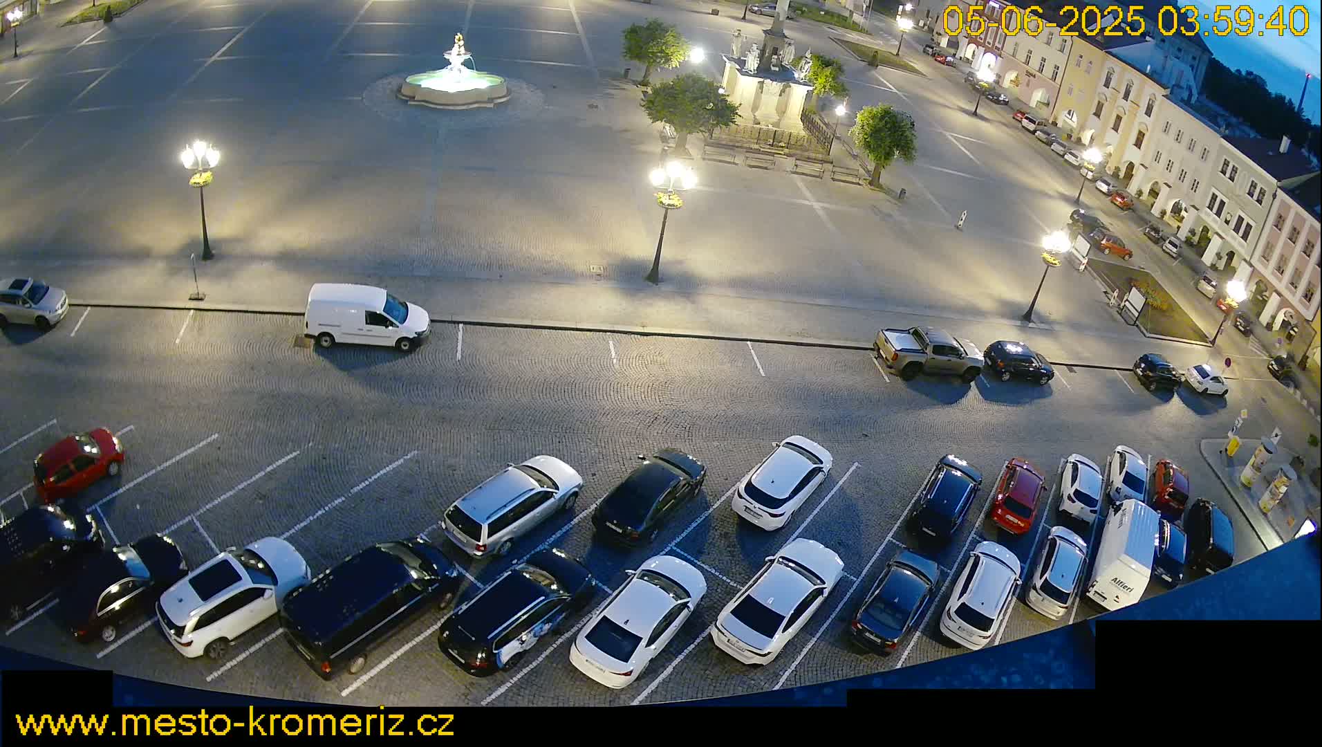 A nighttime, high-angle view of a town square with a fountain and a parking lot filled with cars under artificial light.