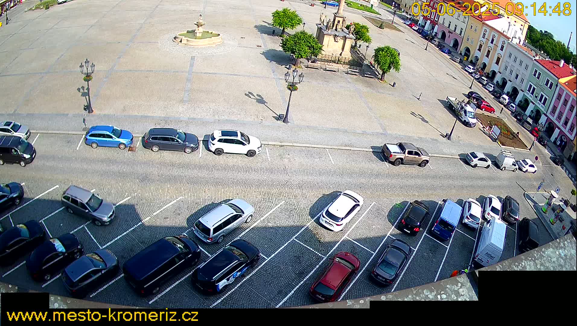 A sunny day shows a town square with a fountain and parked cars lining the surrounding streets.
