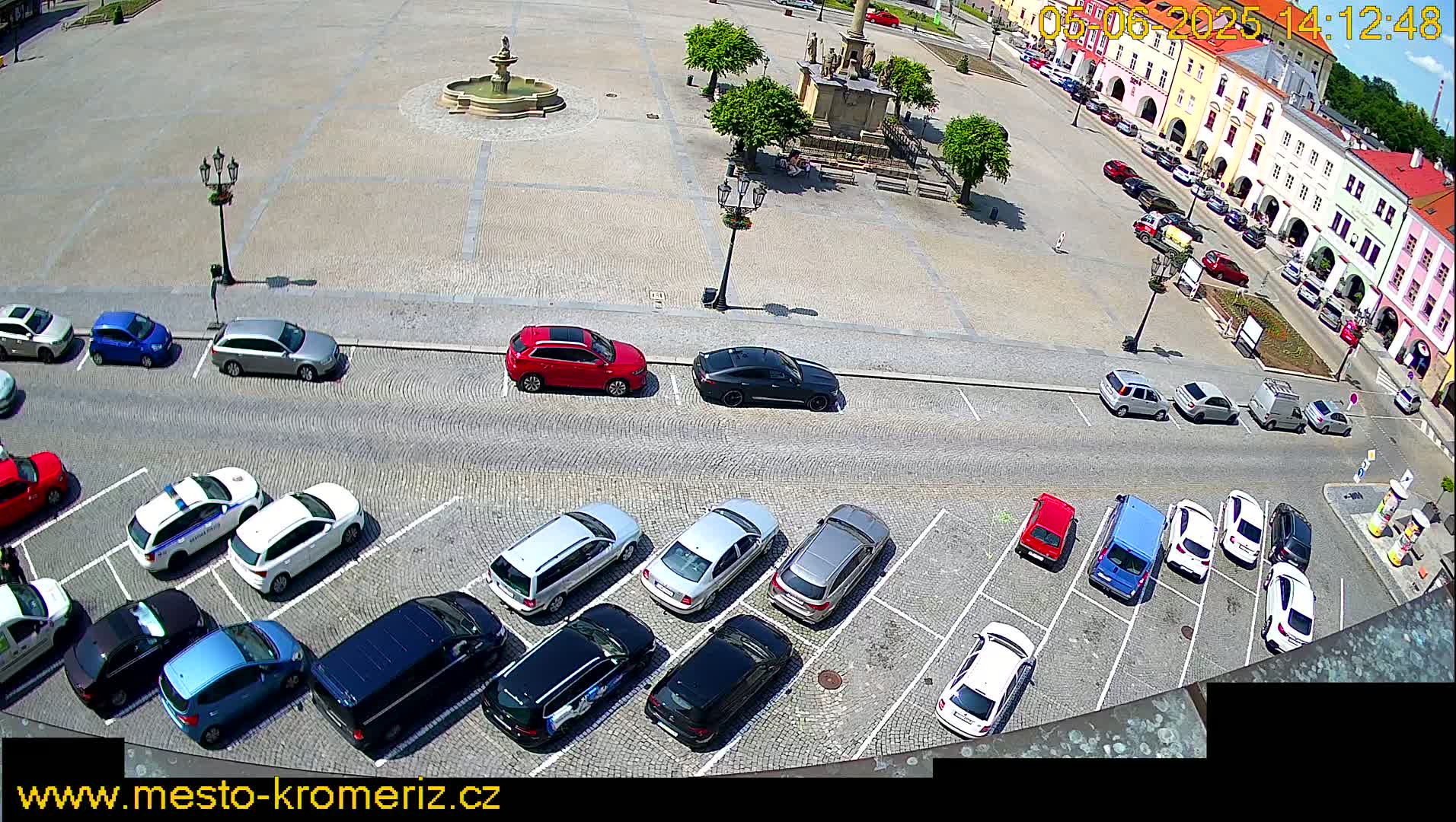 A town square with a fountain is surrounded by parked cars under a sunny sky.