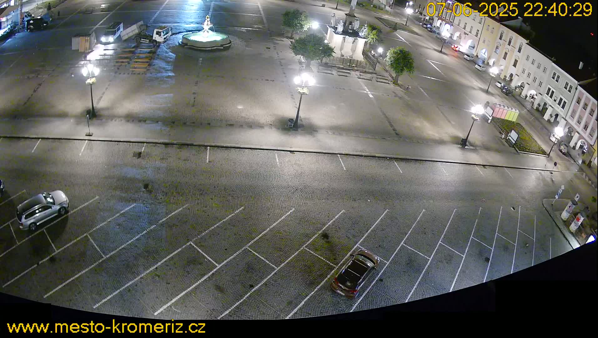 A nighttime, overhead view of a town square with a fountain, parking lot, and several cars.