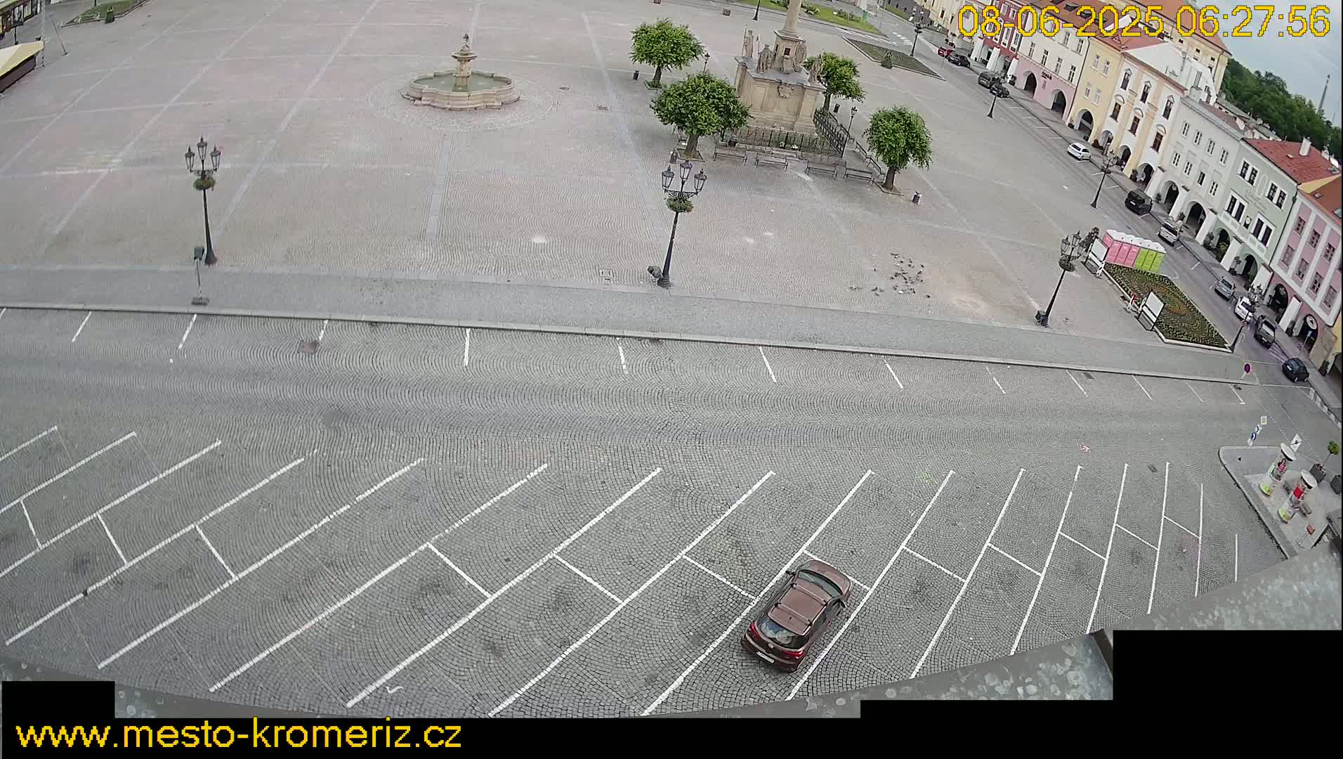 An overhead view shows a mostly empty cobblestone town square with a fountain and a monument, a parking lot, and several buildings surrounding the square on a cloudy day.