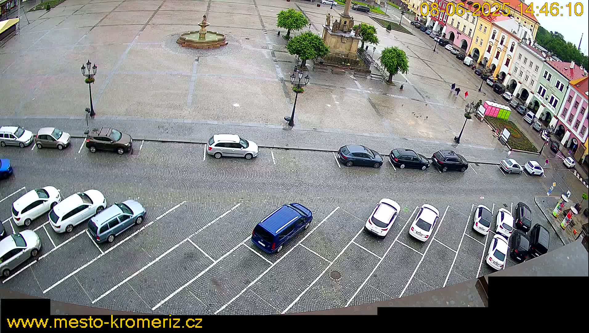 A wet cobblestone town square with a fountain, several parked cars, and a few pedestrians is viewed from above.