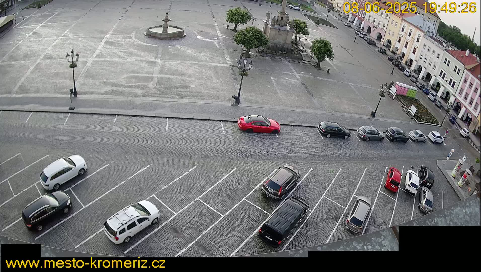 A cobblestone town square with a fountain and several cars parked in a lot adjacent to it, under slightly overcast skies.