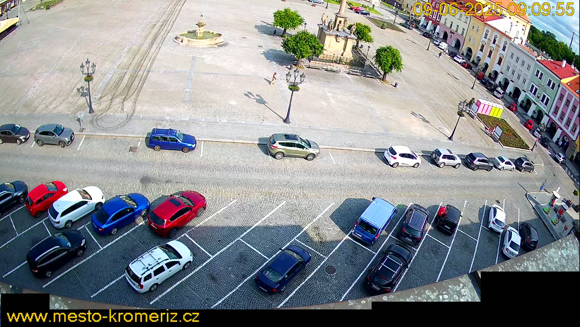 A paved town square with a fountain and several parked cars, under sunny conditions.