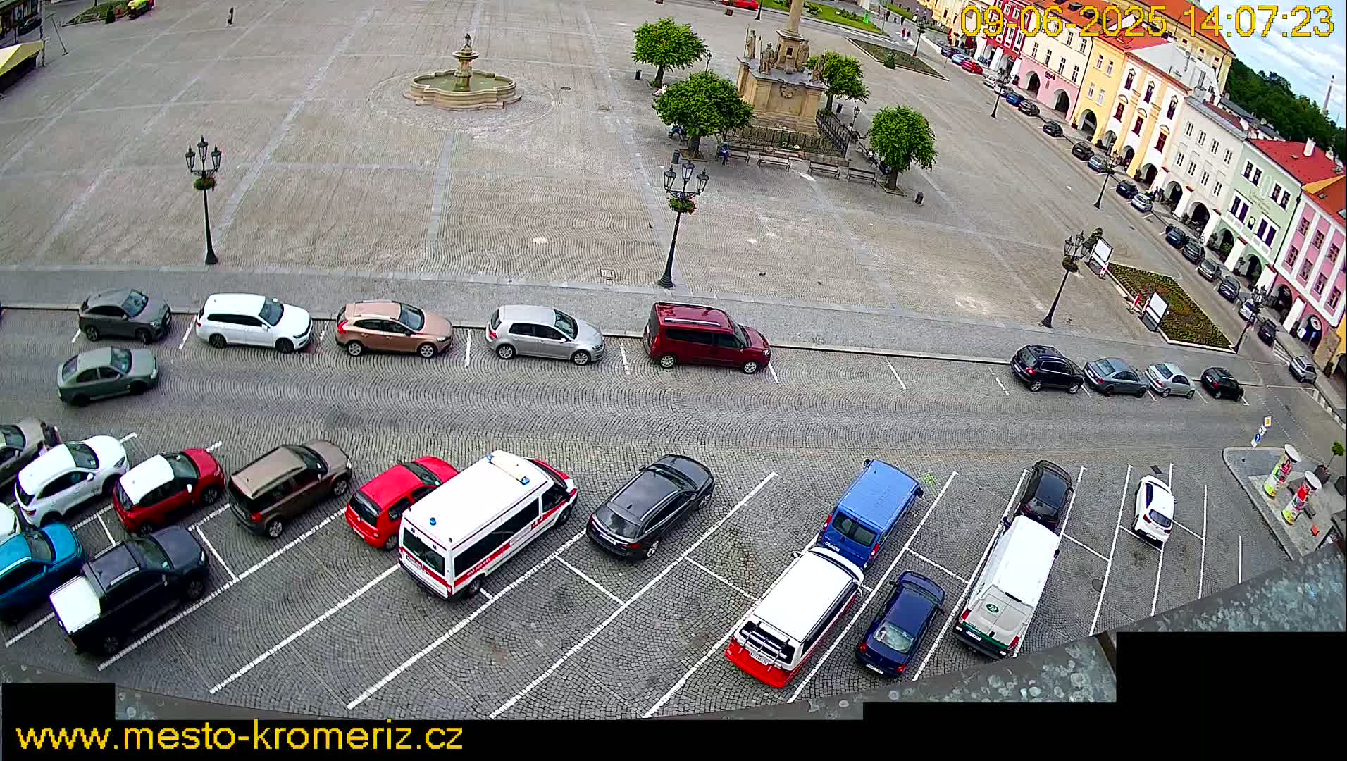 An aerial view shows a cobblestone town square with a fountain and several parked cars surrounding it on a partly cloudy day.