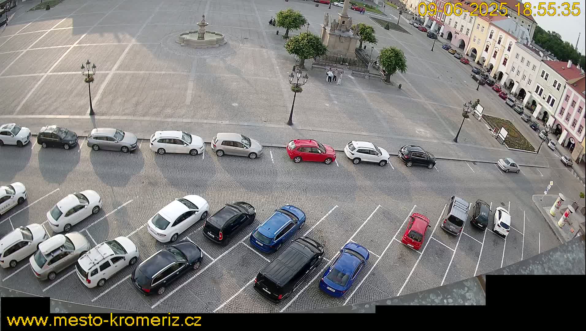 A paved town square with a fountain and a parking lot filled with cars is viewed from above on a sunny day.