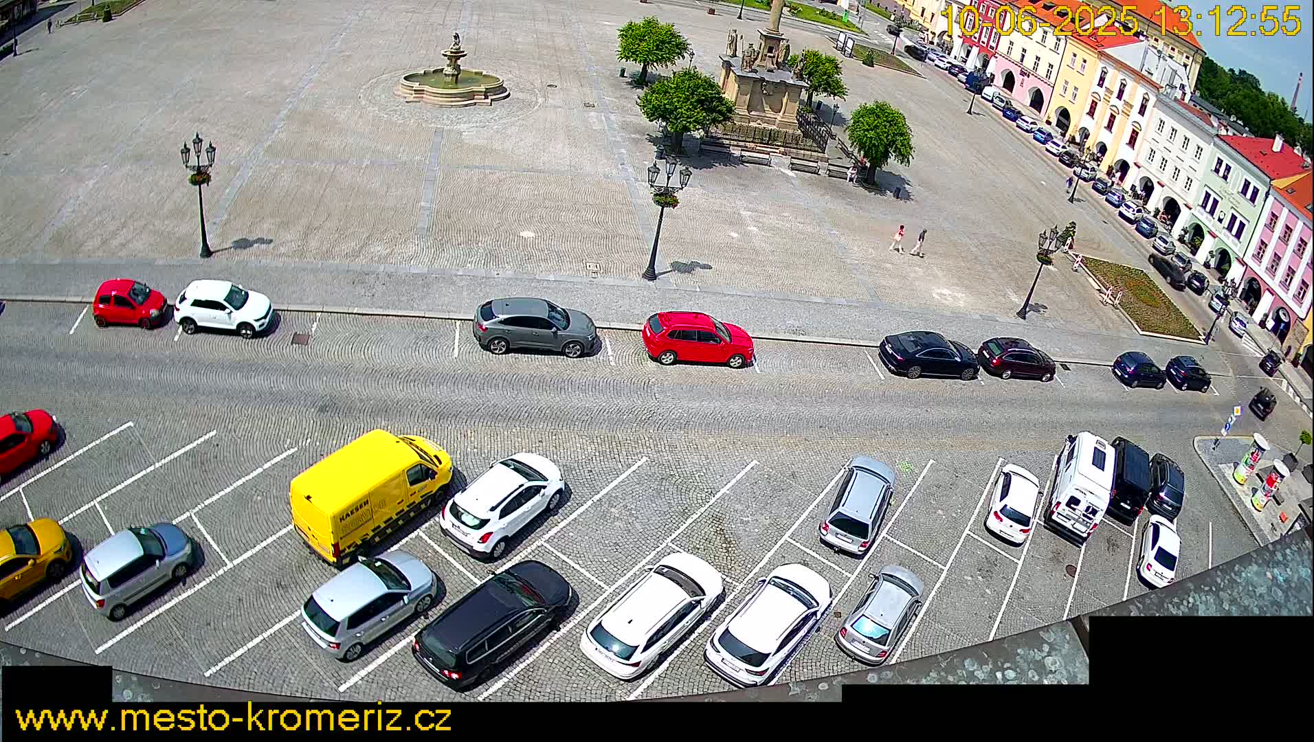 A sunny day shows a town square with several cars parked in a lot, and additional cars driving along the side of the square.