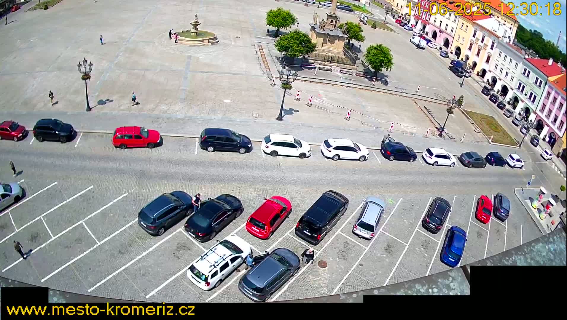 A sunny day reveals a town square with a fountain and surrounding buildings, and a parking lot filled with numerous cars.