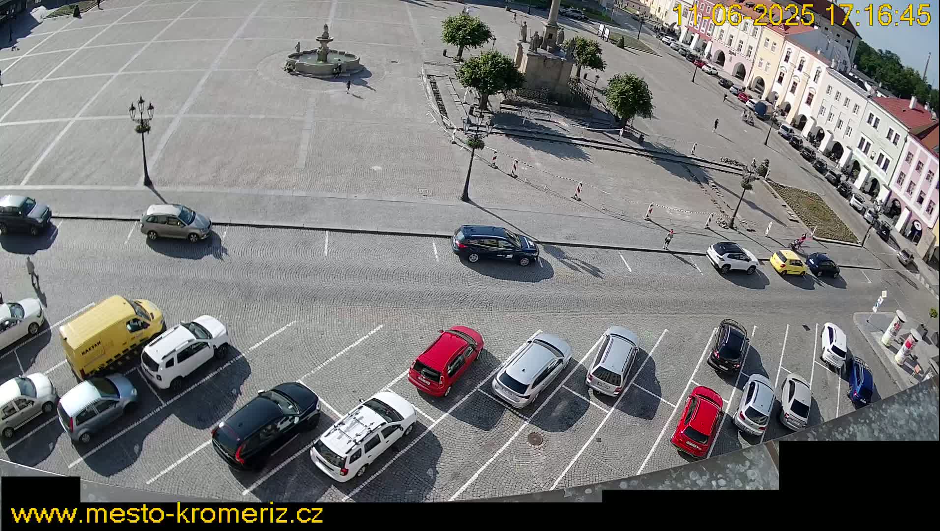 A sunny day reveals a town square with a fountain, surrounded by parked cars and buildings, some of which have arcades.