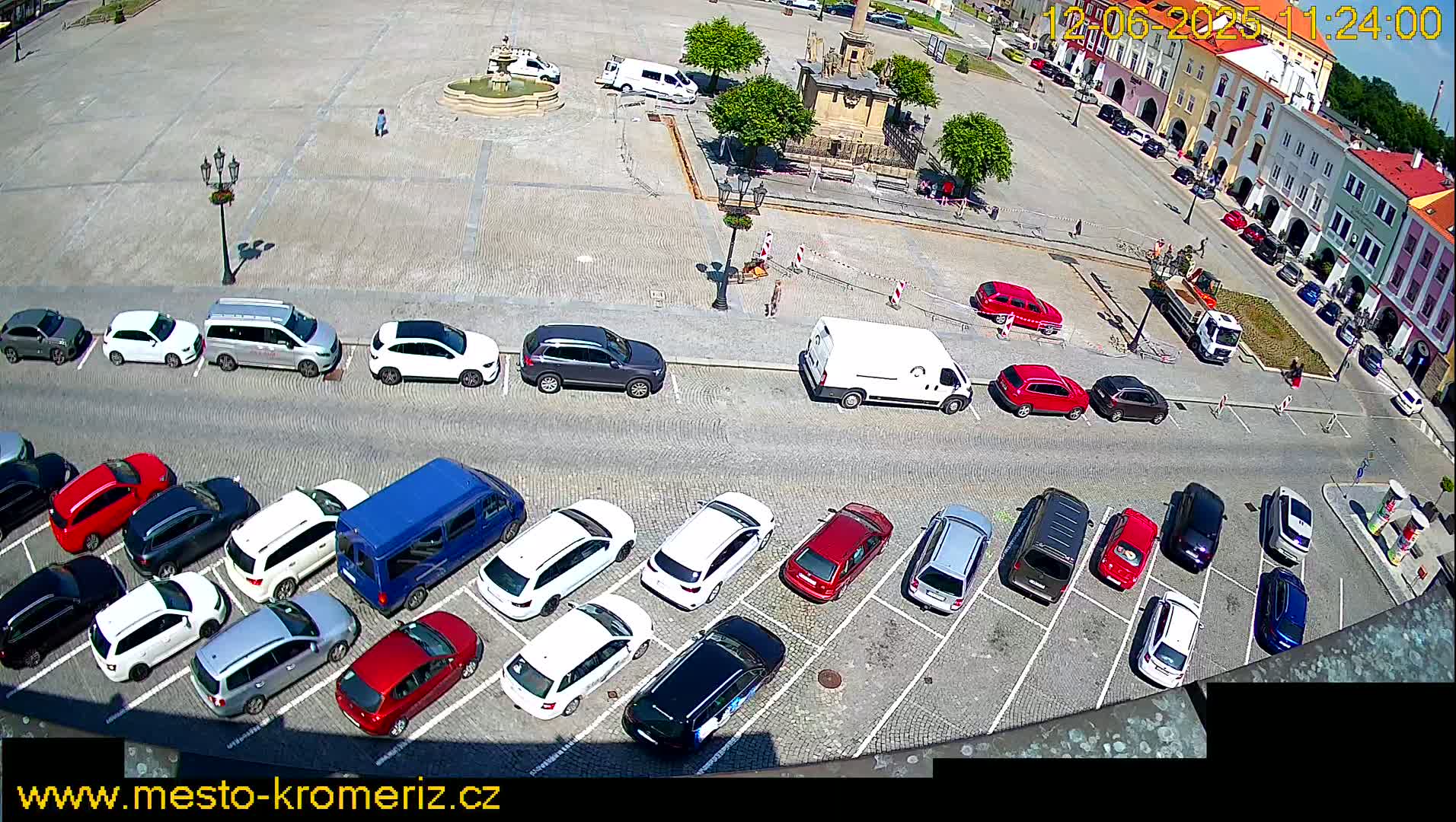 A sunny day shows a town square with a fountain and many cars parked in a lot surrounding the square.