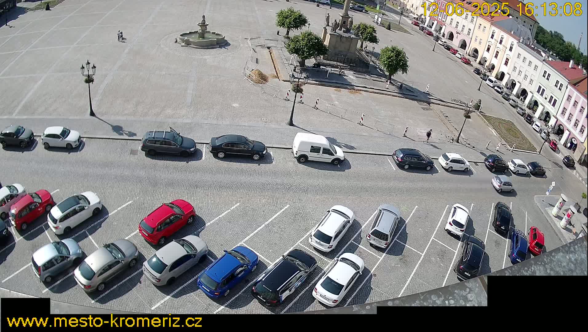 A sunny day in a town square shows a large, paved area with parked cars surrounding a central fountain and monument, with several buildings visible in the background.
