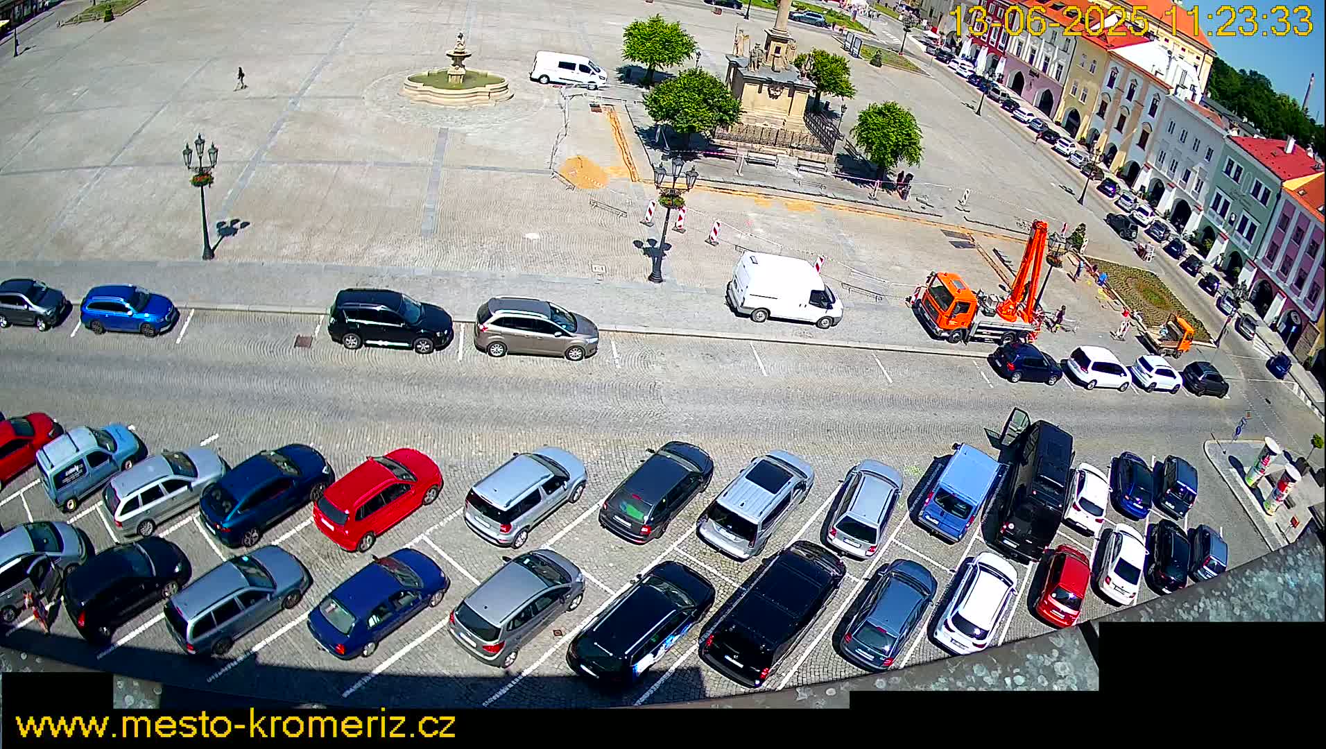 A sunny day shows a town square with a fountain, several parked cars, and construction equipment.