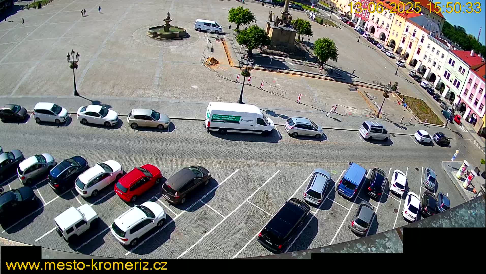 A sunny aerial view of a town square with parked cars, a fountain, and buildings surrounding it.