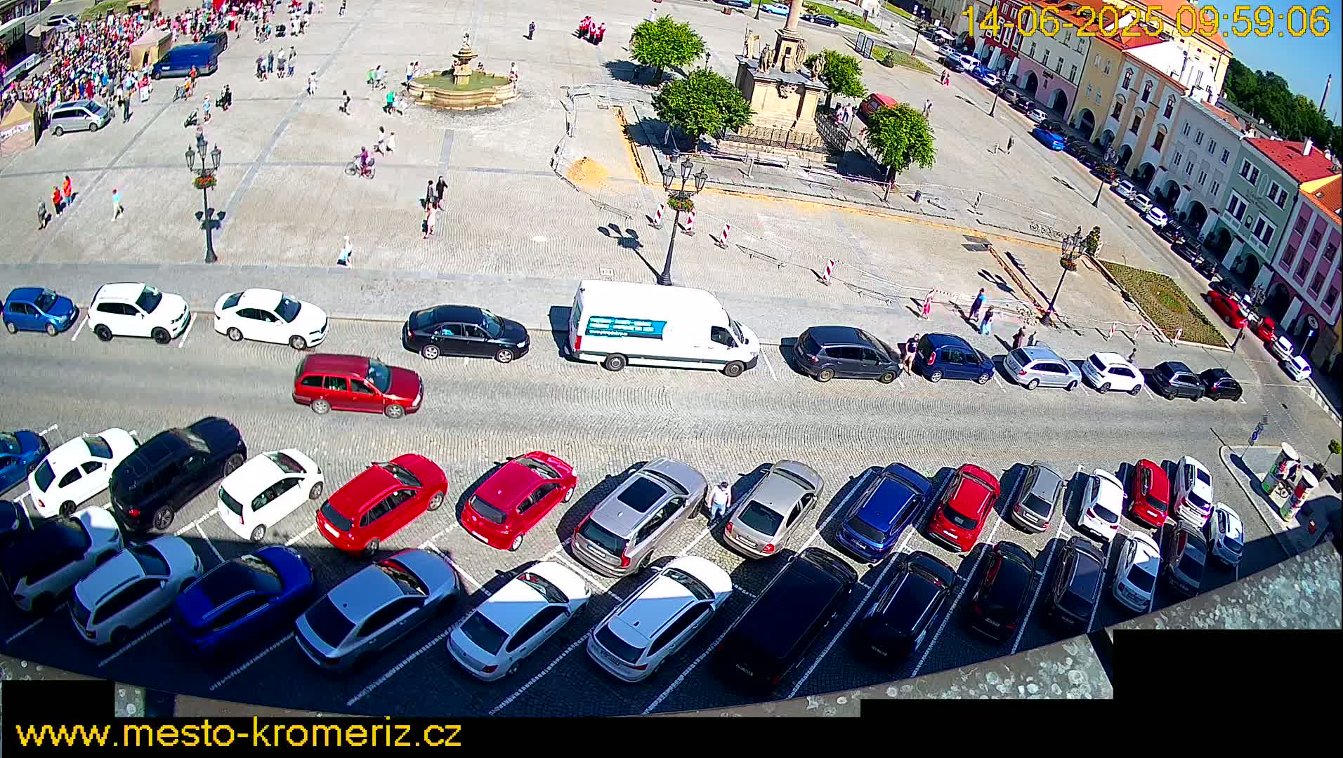 A large town square with a fountain and many parked cars, under sunny conditions.