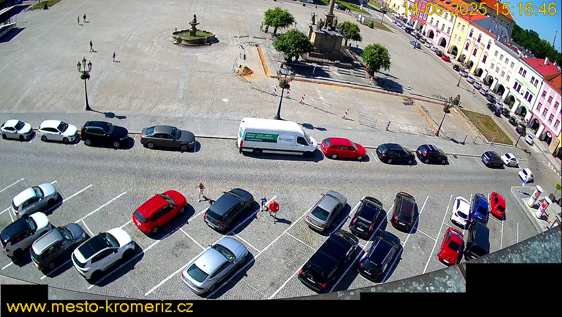 A town square with a fountain, surrounded by buildings and parked cars, on a sunny day.