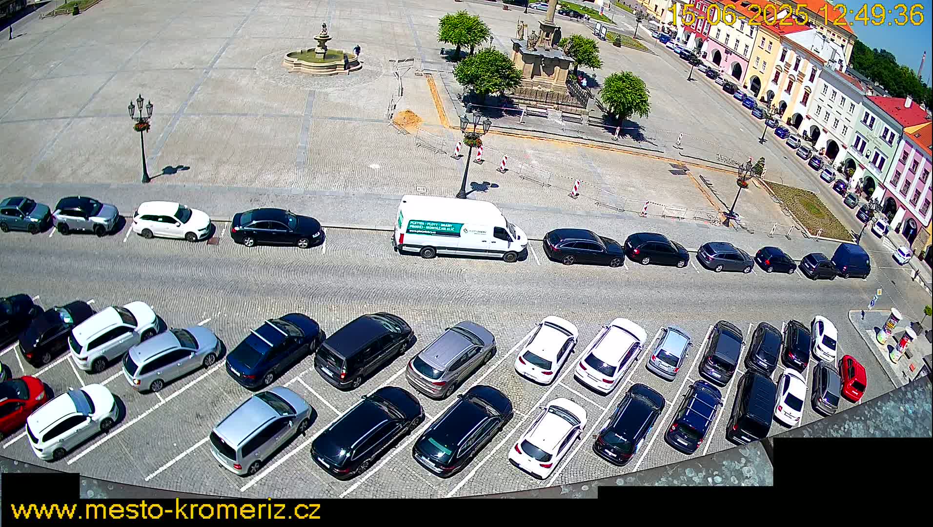 A sunny day shows a large cobblestone square with a fountain and several parked cars, along with a van, surrounding a town square with buildings.