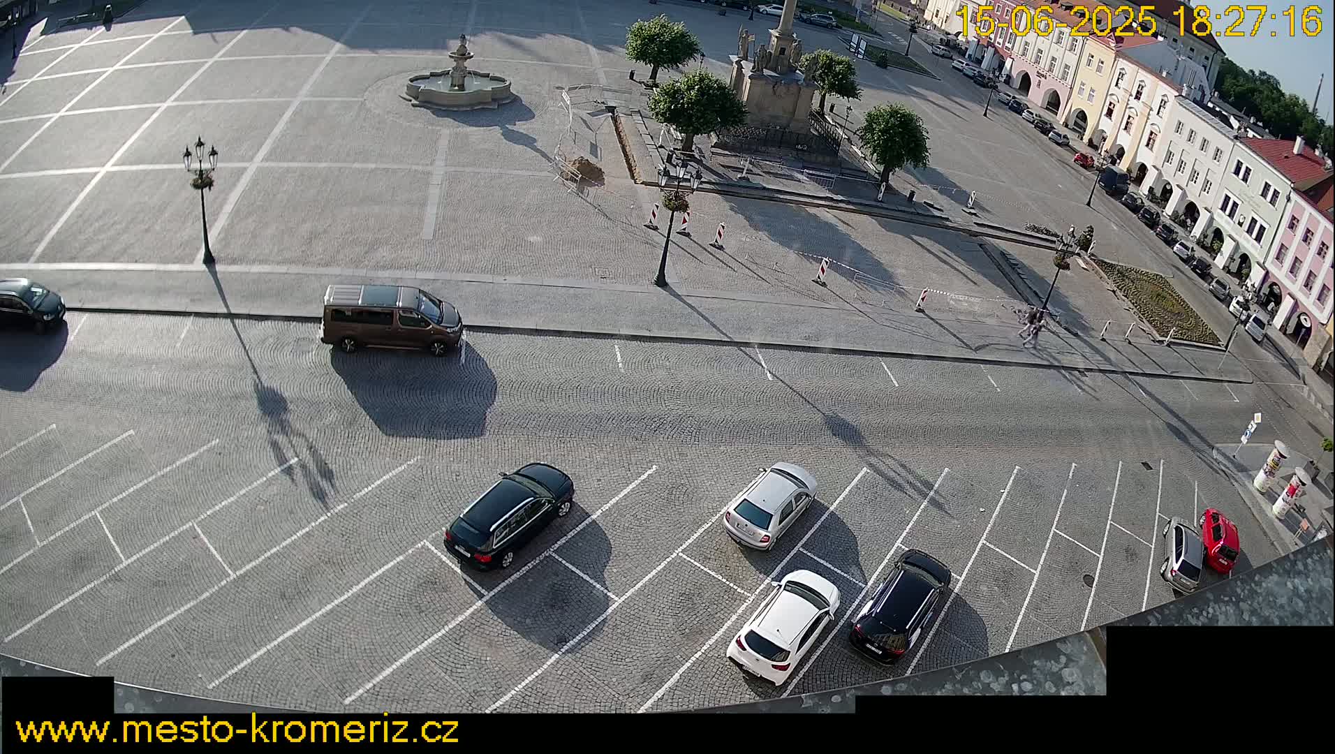 A high-angle, sunny view shows a town square with a fountain and several cars parked in a lot adjacent to a cobblestone street.