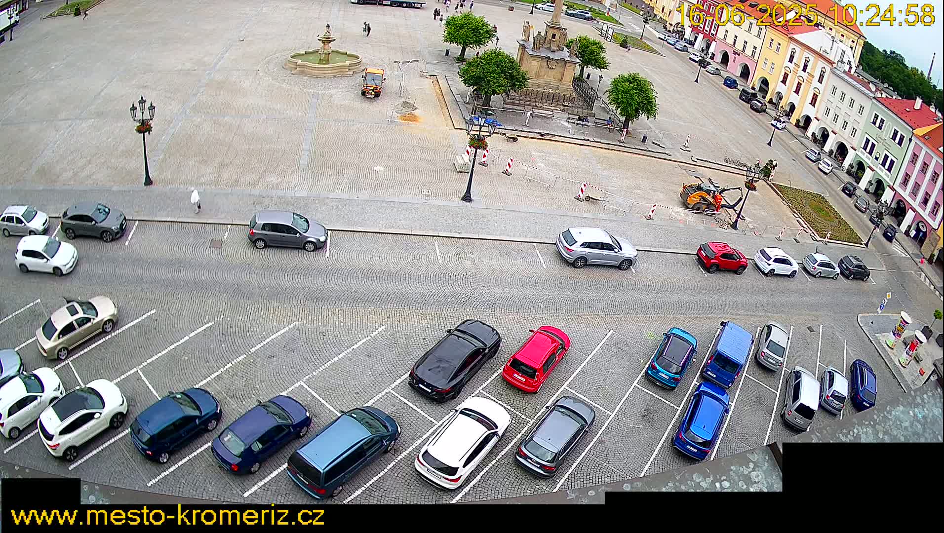 An overcast day shows a town square with a fountain and several cars parked in a lot surrounding it, along with some construction equipment and workers.