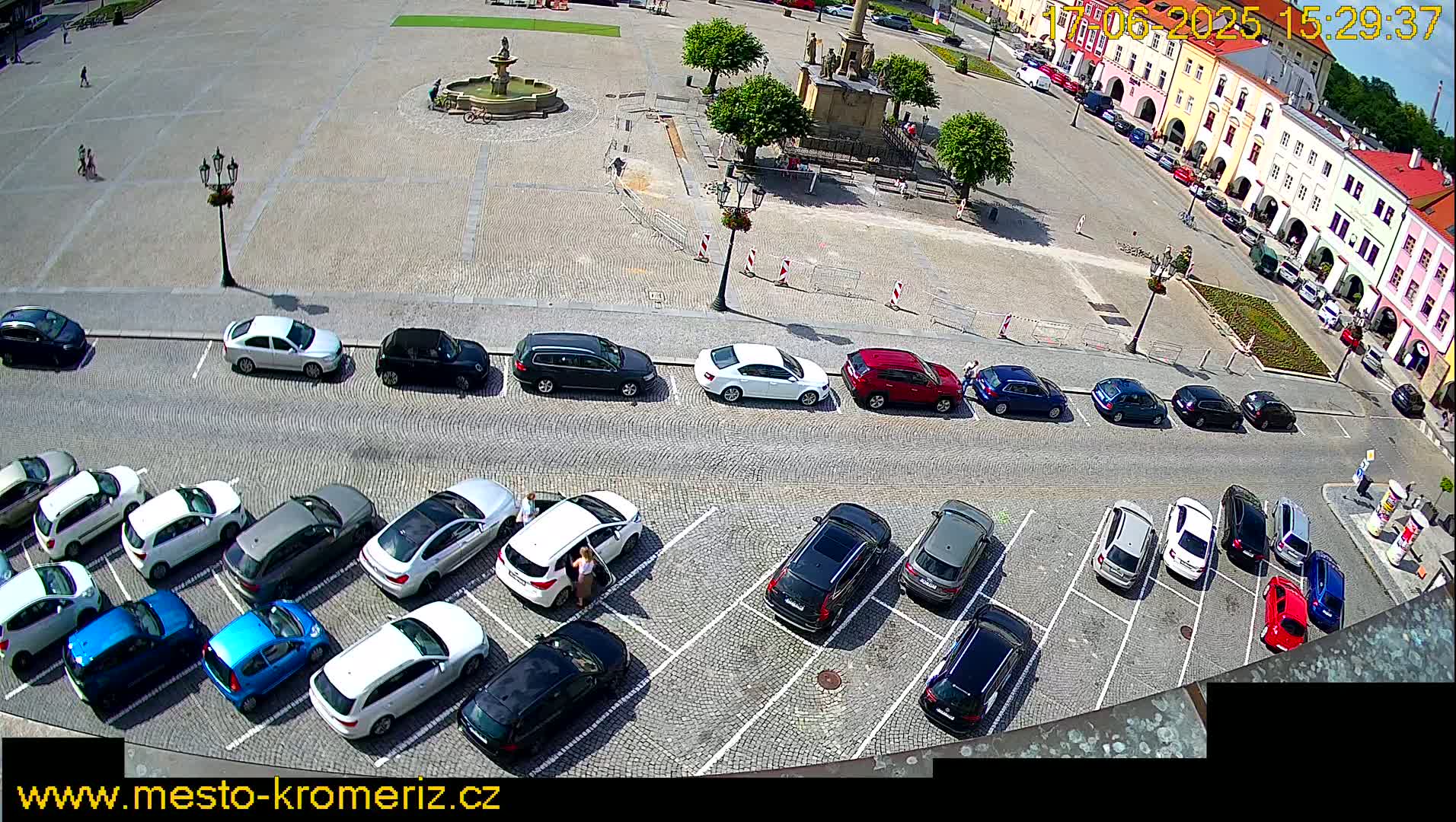 An aerial view of a town square with a fountain, surrounded by buildings and a parking lot filled with cars on a sunny day.