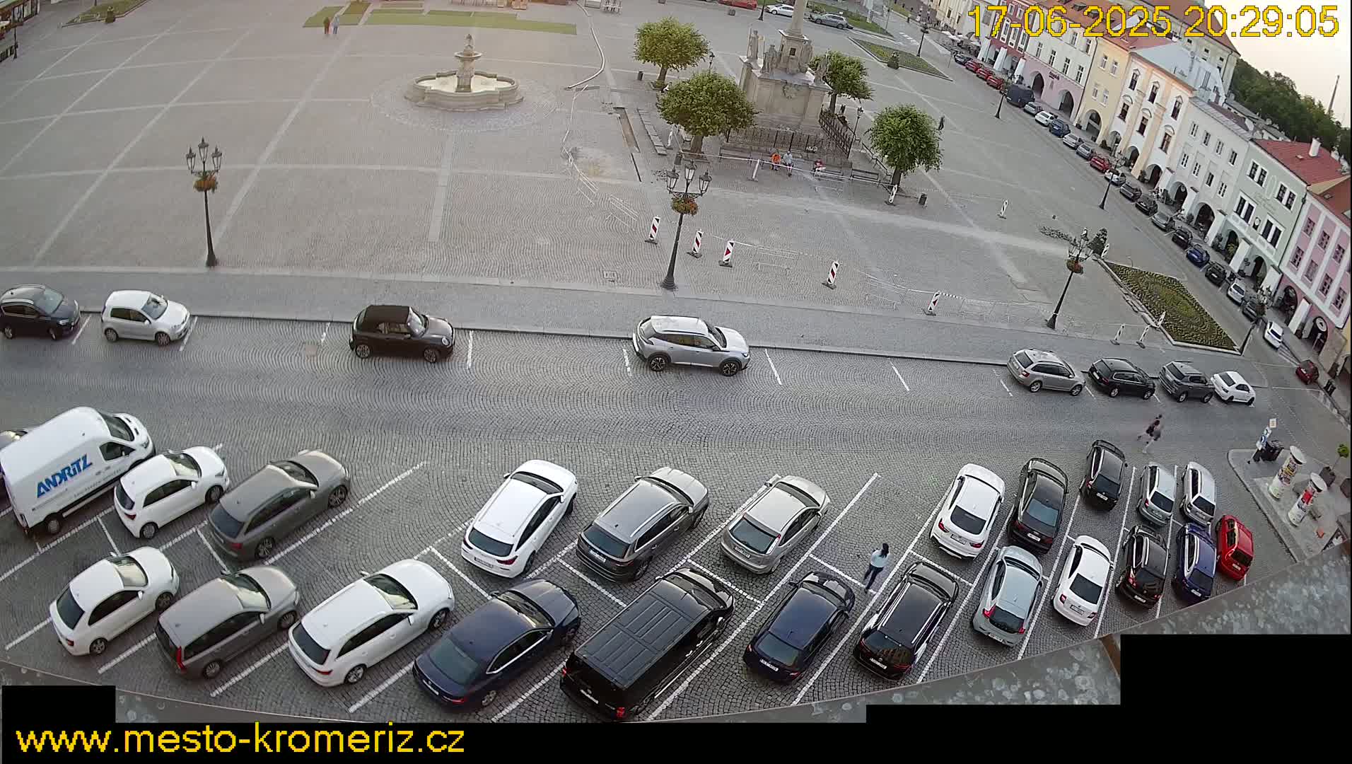 A paved town square with a fountain and several cars parked in a lot beside it, under a twilight sky.